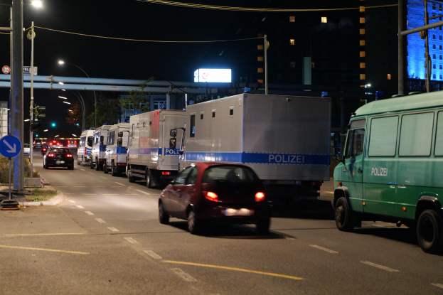 Eine Reihe von Polizeiwagen und Trucks parkt am Straßenrand bei Nacht, mit Gebäuden, einer Brücke und Bäumen im Hintergrund.
