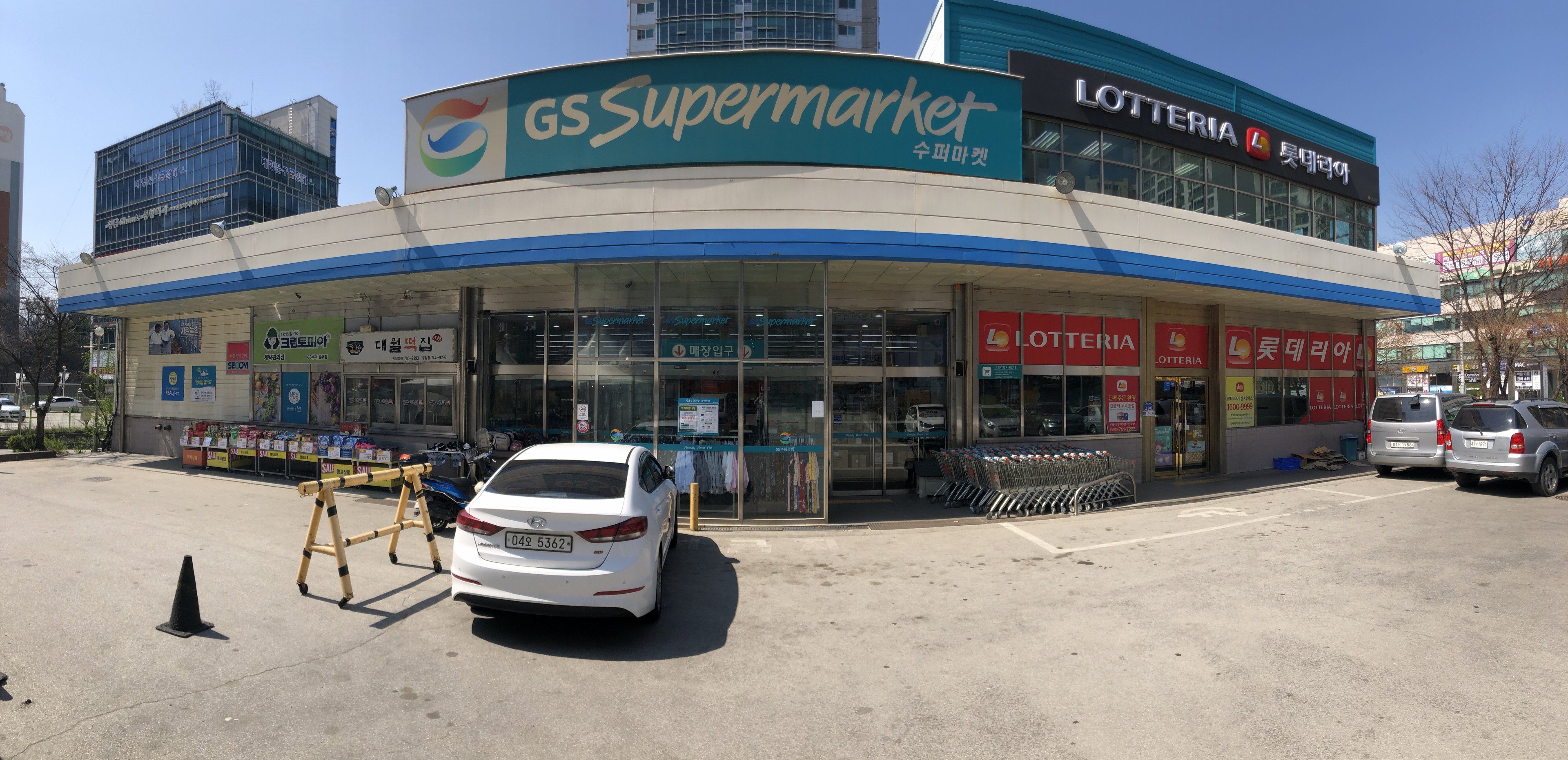 Ein Auto vor einem 'GS Supermarkt' mit anderen Fahrzeugen, Verkehrskegeln und Gegenständen auf der Straße, Gebäuden und Bäumen im Hintergrund bei klarem blauem Himmel.