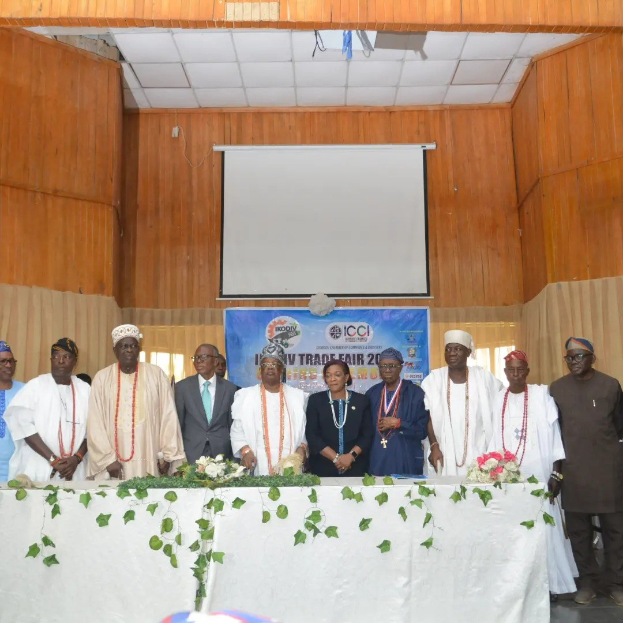 Gruppe von Menschen, die um einen weißen Tisch mit Blumenschmuck auf der ICCI International Trade Fair 2019 stehen, mit einem Banner, Projektionsbildschirm und Deckenbeleuchtung im Hintergrund.