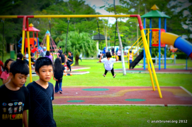Kinder beim Spielen auf Equipment in einem Park mit Bäumen im Hintergrund.