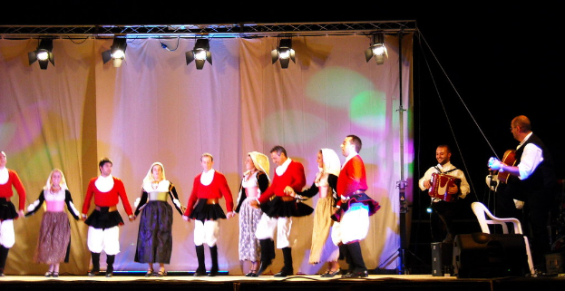 Eine Bühnenperformance mit Menschen auf der Bühne, ein Vorhang im Hintergrund, Lichter oben, Musiker rechts und ein weißer Stuhl unten rechts.