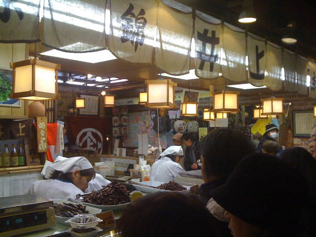 Mitarbeiter arbeiten hinter einer Theke in einem Restaurant mit Gegenständen und Gästen im Vordergrund und Soßenflaschen, Menükarten, Kleidung und Lampen im Hintergrund.