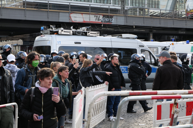 Eine Gruppe von Demonstranten steht vor Polizeifahrzeugen, einige tragen Helme und filmen mit Handys, während Beamte versuchen, sie in der Nähe von Barrikaden, einer Brücke und Gebäuden in Berlin zu zerstreuen.