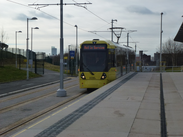 Eine Straßenbahn auf ihren Schienen mit Straßenlaternen, Strommasten, einem Eisengitter und einem Bahnsteig, mit Gebäuden, Bäumen und dem Himmel im Hintergrund.