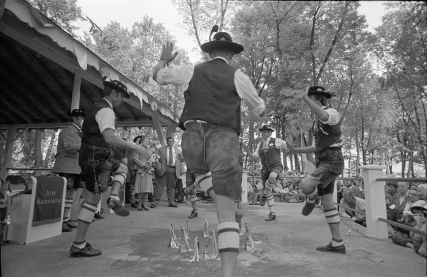 Eine Gruppe von Männern in traditioneller bayrischer Tracht tanzt auf einer Bühne vor einem sitzenden und stehenden Publikum, mit Bäumen und einer Hütte im Hintergrund, dargestellt in Schwarz-Weiß.