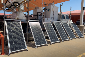 Eine Gruppe von Solarmodulen auf einem Parkplatz mit Fahrzeugen, Pfählen, Lampen und anderen Gegenständen sowie Hütten und einem klaren blauen Himmel im Hintergrund.