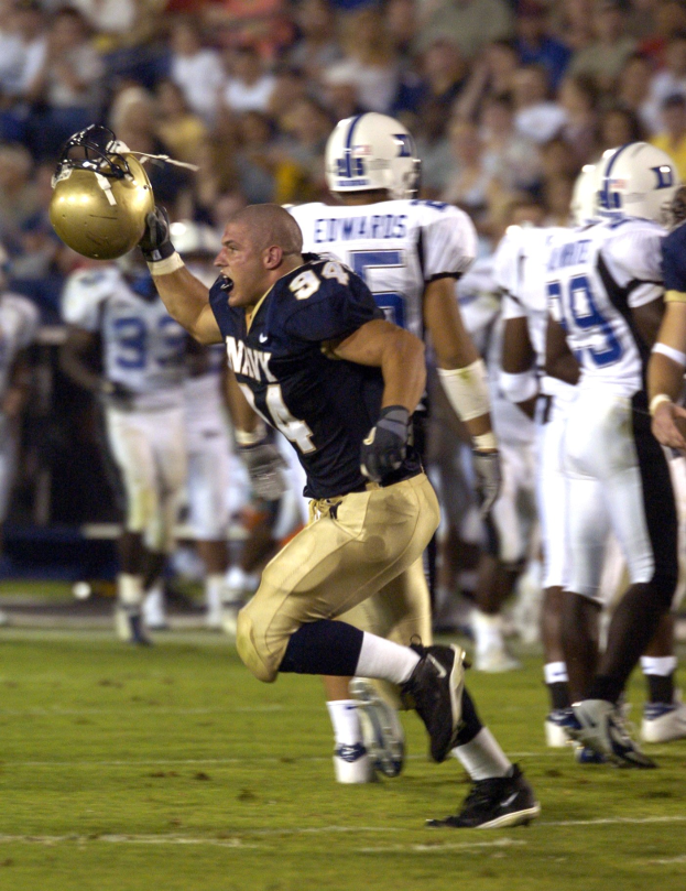 Ein Fußballspieler in dunkler Uniform rennt mit dem Ball, umgeben von Teamkollegen und Gegnern in Helmen und Sportbekleidung auf dem Feld, während Zuschauer auf Stühlen im Hintergrund sitzen.
