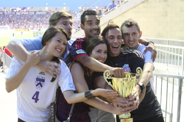 Sechs Personen, zwei Frauen und vier Männer, die einen Pokal halten und lächeln, posieren für ein Foto vor einem Eisenzaun, mit einer Wand und einem Zaun oben rechts, Menschen und Lichter im Hintergrund, möglicherweise in einem Stadion.