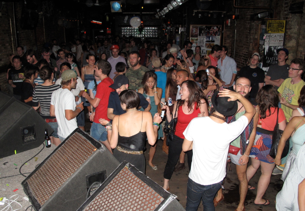 Eine Gruppe von Menschen, die tanzen und einige Mützen tragen und Taschen tragen, mit einer Soundbox, Kabeln, einer Flasche und Texttafeln im Hintergrund.