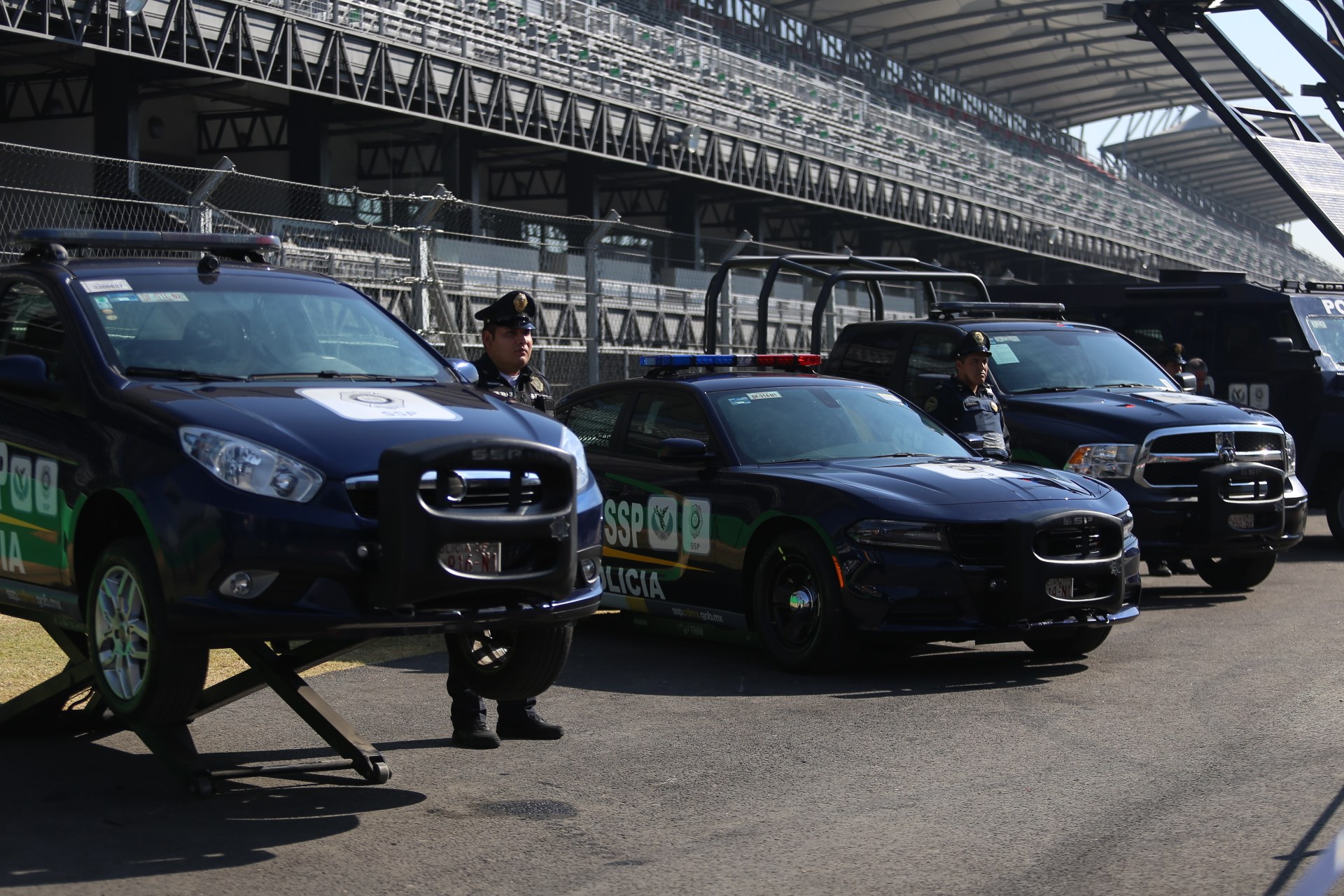 Polizeiautos vor einer Rennstrecke mit uniformierten Personen, im Hintergrund ein Stadion mit Gittern und Maschendraht.