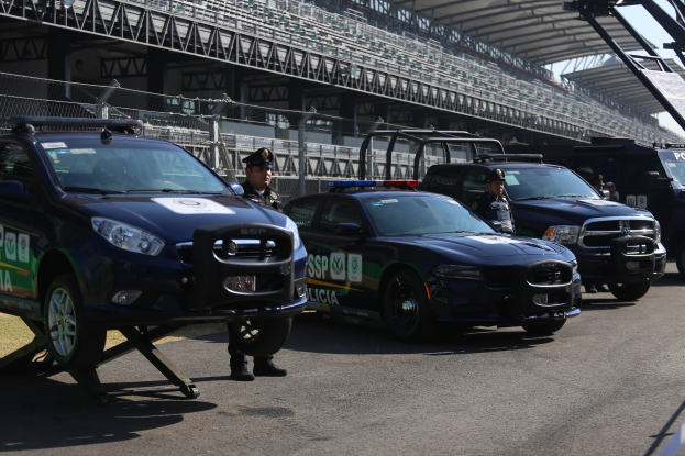 Polizeiautos vor einer Rennstrecke mit uniformierten Personen, im Hintergrund ein Stadion mit Gittern und Maschendraht.