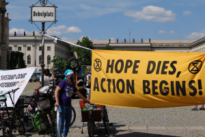 Eine Gruppe von Menschen steht vor einem Gebäude und hält ein gelbes Banner mit der Aufschrift "Hope Dies, Action Begins". In der Nähe stehen mehrere Fahrräder und ein Pfahl mit einer Tafel daran. Im Hintergrund sind Bäume und ein klarer blauer Himmel zu sehen.
