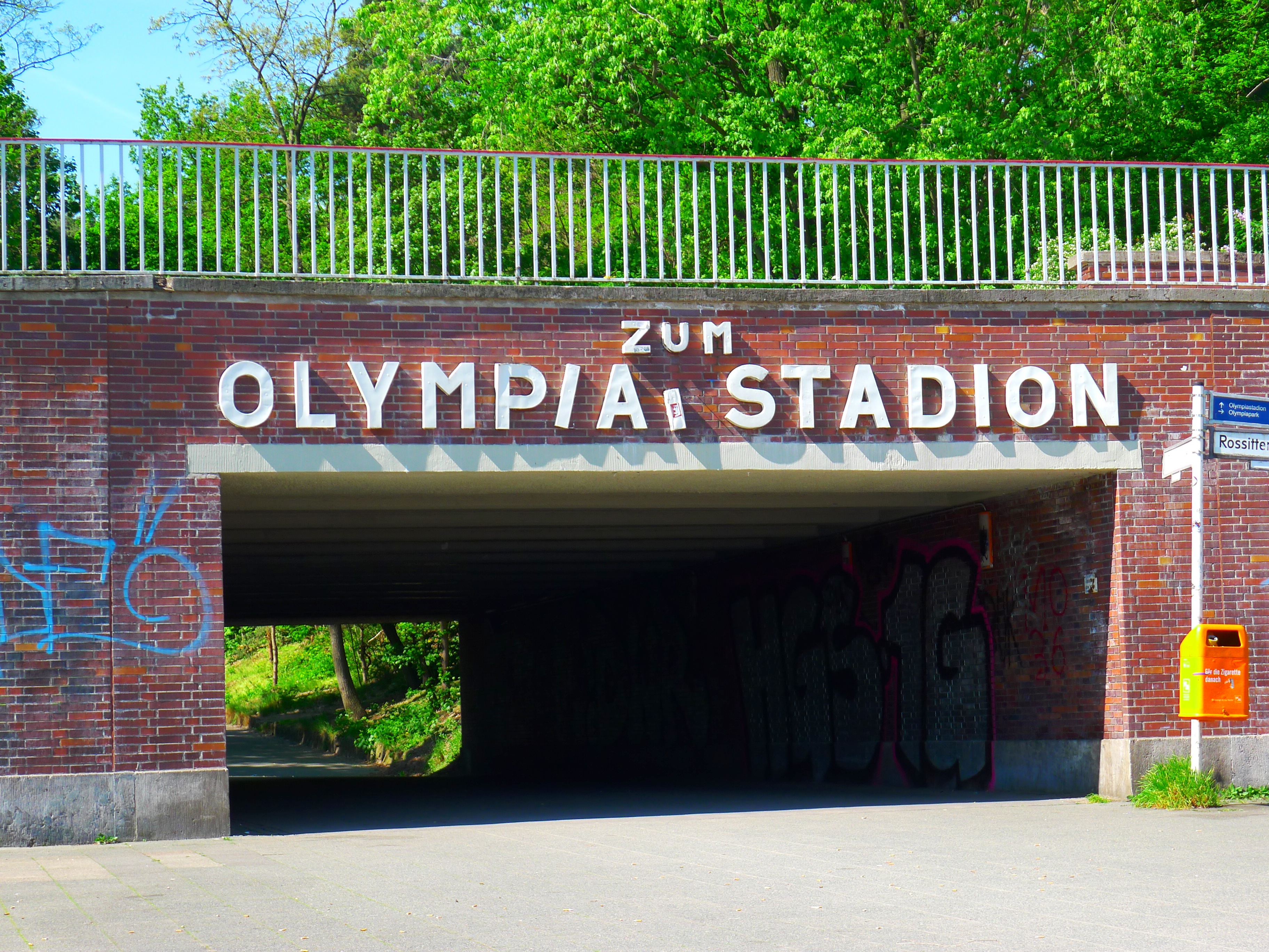 Der Eingang zum Olympiastadion in Berlin, Deutschland, mit einer Brücke mit Text, einem Metallzaun, einer Tafel, einem Kasten, Pflanzen, Gras, einer Baumgruppe und einem bewölkten Himmel.