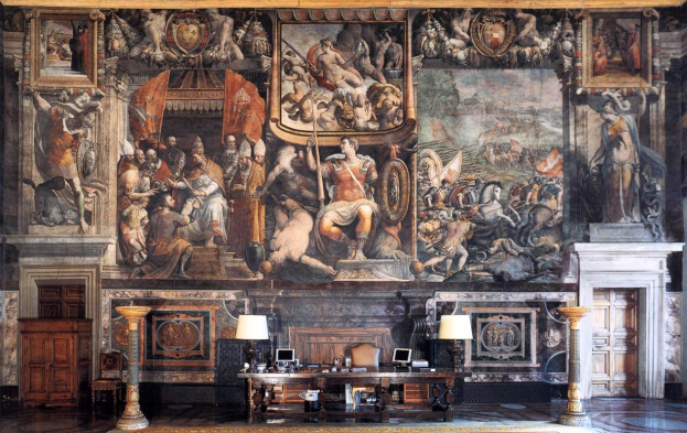 Ein geräumiger Raum im Palazzo della Signoria in Florenz, Italien, mit einem großen Gemälde, einem Tisch mit Gegenständen, zwei Lampen, zwei Türen und einem Teppich.