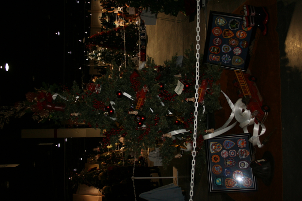 Ein Weihnachtsbaum mit Geschenken, Rahmen und Spielzeugen auf einer Plattform, mit Menschen, weiteren Weihnachtsb├Ąumen, einer Kette, einem Pfosten und Lichtern im Hintergrund.