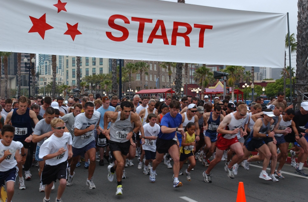 Gruppe von Menschen beim Marathon mit einem Verkehrskegel im Vordergrund und einem Textbanner im Hintergrund, vor einer Kulisse aus Bäumen, Laternenmasten, Gebäuden und einem klaren blauen Himmel.