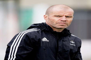 Ein kahlköpfiger Trainer in einer blauen Jacke mit weißen Streifen und weißem Kragen steht auf einem Fußballfeld mit gefalteten Händen und ernster Miene.