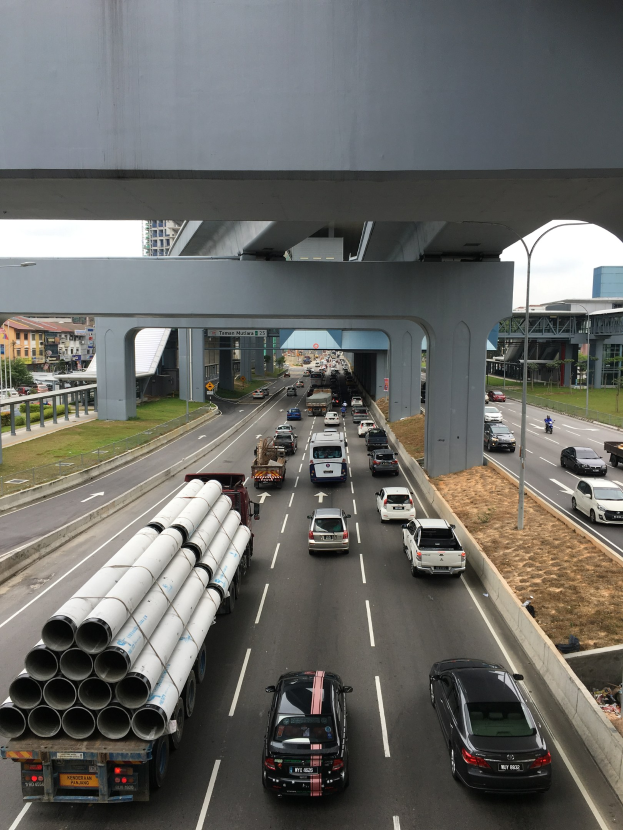 Eine vielbefahrene Autobahn mit mehreren Fahrzeugen, einer Brücke darüber, Straßenlaternen, Gras, Gebäuden, Bäumen und einem Himmel als Hintergrund.