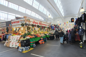 Ein belebter Indoor-Markt mit Menschen, die zwischen Ständen mit Obst, Gemüse und aufgehängten Kleidern unter Deckenlampen gehen, vor hellen Glasfenstern.