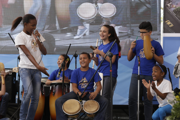 Gruppe von Kindern, die auf einer Bühne Musikinstrumente spielen, mit einer Frau in der Mitte, die ein Mikrofon hält, umgeben von Mikrofonstativen und einem Banner im Hintergrund, mit Pflanzen auf der rechten Seite.