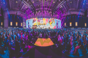 Eine große Menschenmenge, die auf Stühlen vor einer hell erleuchteten Bühne sitzt, mit mehreren stehenden Personen, wahrscheinlich auf einem Online-Marketing-Rockstars-Event, umgeben von Bildschirmen.