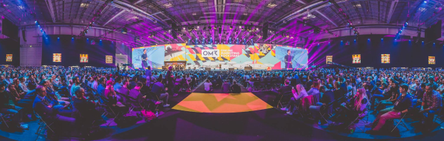 Eine große Menschenmenge, die auf Stühlen vor einer hell erleuchteten Bühne sitzt, mit mehreren stehenden Personen, wahrscheinlich auf einem Online-Marketing-Rockstars-Event, umgeben von Bildschirmen.