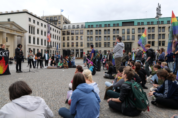 Eine Gruppe von Menschen, die auf dem Boden vor einer Menge sitzt, die Fahnen und Transparente hält, während einer Anti-Schwulen-Demo in Berlin, mit einer Statue, Gebäuden und einem bewölktem Himmel im Hintergrund.