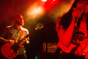 Eine Frau singt in ein Mikrofon auf der Bühne, wür ein Mann eine Akustikgitarre im Hintergrund spielt, mit Deckenlampen, die die lebendige Szenerie beleuchten.