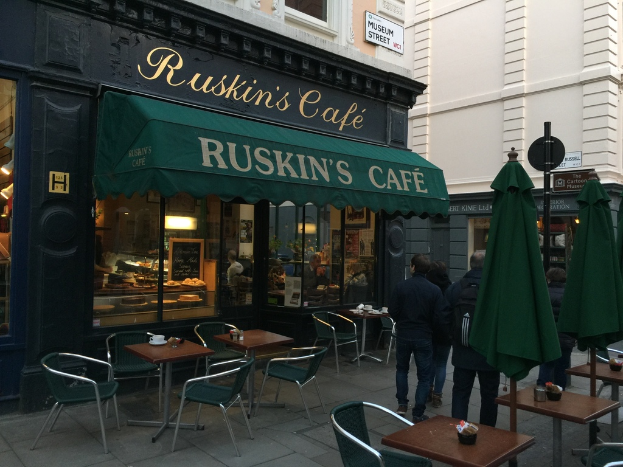 Eine Gruppe von Menschen steht vor einem Cafe mit einem Schild, auf dem "Ruskin's Cafe" steht.