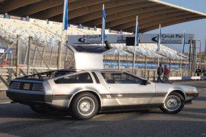 Ein silberner DeLorean DMC-12 Wagen befindet sich auf einer Straße umgeben von Menschen, Fahnen, Schildern, Geländern, Stühlen und einem Schuppen, mit Bäumen und einem klaren blauen Himmel im Hintergrund.