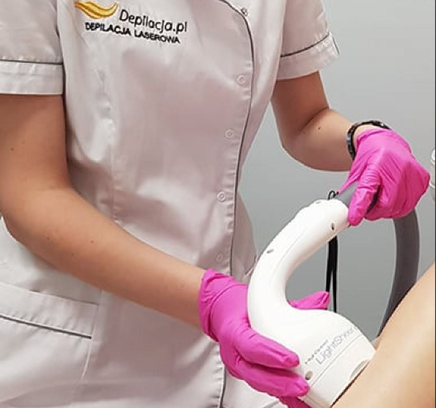 Eine Frau in einem weißen Hemd und pinken Handschuhen bedient eine weiße Laser-Haarentfernungsmaschine auf ihrer Haut, während sie vor einer Wand steht.