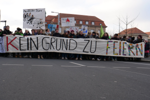Gruppe von Menschen mit einem Transparent mit der Aufschrift "Kein Grund zu Feiern" bei einer Demonstration gegen die Sparpolitik der deutschen Regierung, im Hintergrund sind Straßenlaternen, Bäume, Gebäude mit Fenstern und ein klarer Himmel zu sehen.