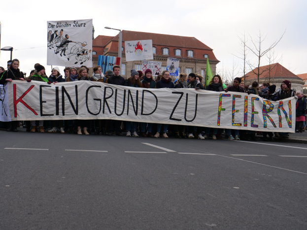 Gruppe von Menschen mit einem Transparent mit der Aufschrift "Kein Grund zu Feiern" bei einer Demonstration gegen die Sparpolitik der deutschen Regierung, im Hintergrund sind Straßenlaternen, Bäume, Gebäude mit Fenstern und ein klarer Himmel zu sehen.