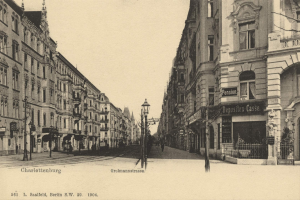 Schwarzes und weißes Foto einer Straße in Charlottenburg, Deutschland, mit Gebäuden, Straßenmasten, Lichtern, Namensschildern, Kraftfahrzeugen, Füßgängern, Gittern und einem klaren Himmel.