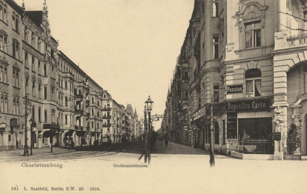 Schwarzes und weißes Foto einer Straße in Charlottenburg, Deutschland, mit Gebäuden, Straßenmasten, Lichtern, Namensschildern, Kraftfahrzeugen, Füßgängern, Gittern und einem klaren Himmel.