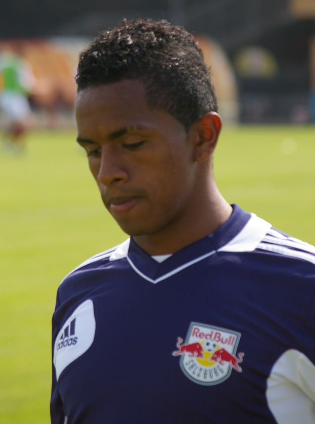 Junger Mann in blauer T-Shirt auf einem Fuballfeld, der als Flügelspieler mit kurzen dunklen Haaren und entschlossener Miene erscheint.