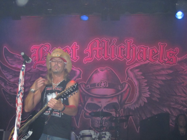 Eine Musikkonzertszene mit einem Gitarristen auf der Bühne, einer Person in der unteren rechten Ecke, einem Banner im Hintergrund und Drogen in der Mitte unten.