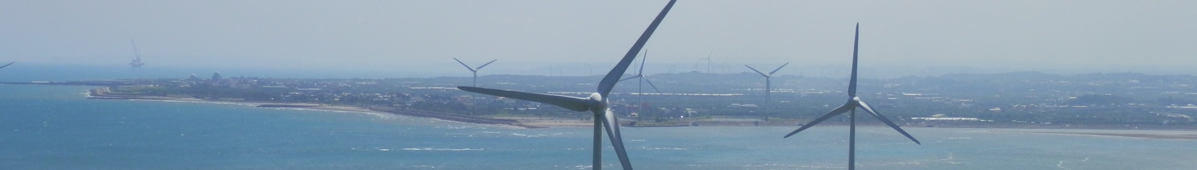 Gruppe von Windkraftanlagen im Meer in der Nähe einer Stadt, mit Wasser, das gegen den Strand schwappt, Bäumen und Gebäuden im Hintergrund und einem klaren blauen Himmel darüber.