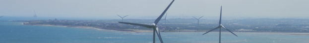 Gruppe von Windkraftanlagen im Meer in der Nähe einer Stadt, mit Wasser, das gegen den Strand schwappt, Bäumen und Gebäuden im Hintergrund und einem klaren blauen Himmel darüber.