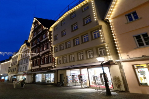 Eine festlich geschmückte Kopfsteinpflasterstraße in Rothenburg ob der Tauber, Deutschland, gesäumt von dekorierten Gebäuden und beleuchtet von Weihnachtslichtern, mit Menschen, die vorbeigehen, und einem Fahrrad in der Nähe.