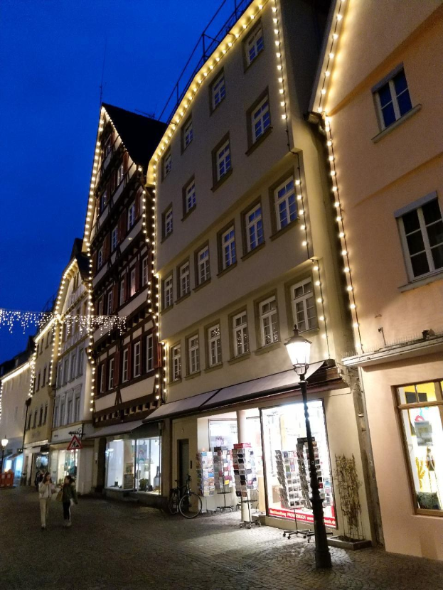 Eine festlich geschmückte Kopfsteinpflasterstraße in Rothenburg ob der Tauber, Deutschland, gesäumt von dekorierten Gebäuden und beleuchtet von Weihnachtslichtern, mit Menschen, die vorbeigehen, und einem Fahrrad in der Nähe.