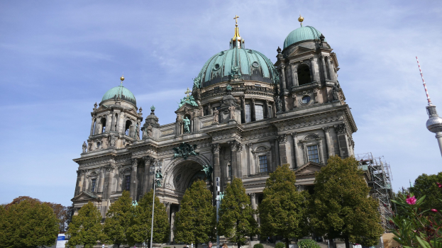 Großes, prunkvolles Berliner Dom-Gebäude in Berlin, Deutschland, mit architektonischen Details wie Säulen, Bögen und Statuen, vor einem bewölkten Himmel, mit Menschen, Bäumen und einem Turm im Vordergrund.