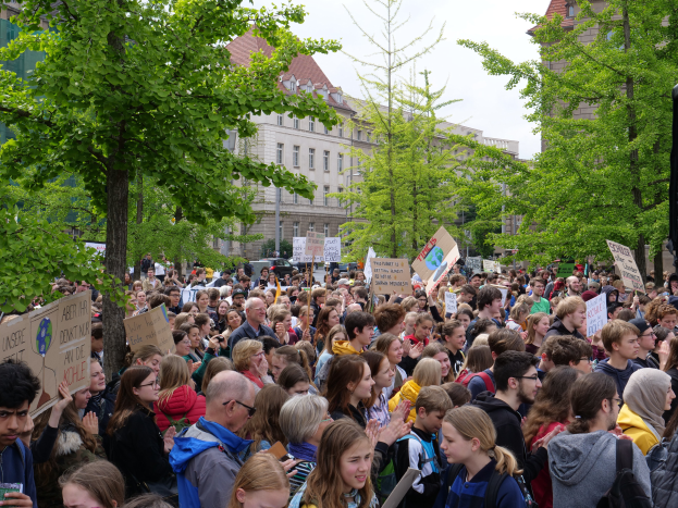 Eine Menschenmenge mit Schildern steht vor einem Gebäude in Berlin, mit Bäumen, Fahrzeugen und einem Redner im Hintergrund.