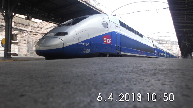 Ein Zug steht im Bild mit einem Bahnsteig und Text unten, Gebäuden und einem Dach links und Gebäuden, Dächern, Kabeln und Himmel oben.