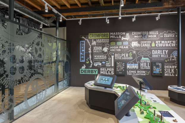 Ein Museumsraum mit zahlreichen Tischen und Monitoren, eine text- und bildbedeckte Wand, eine beleuchtete Decke und eine Glaswand, wahrscheinlich das Cromford Mills Museum.