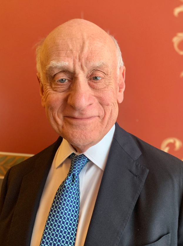 Ein älterer Mann im Anzug und Krawatte lächelt vor einer roten Wand und sieht aus wie ein Nobelpreisträger.