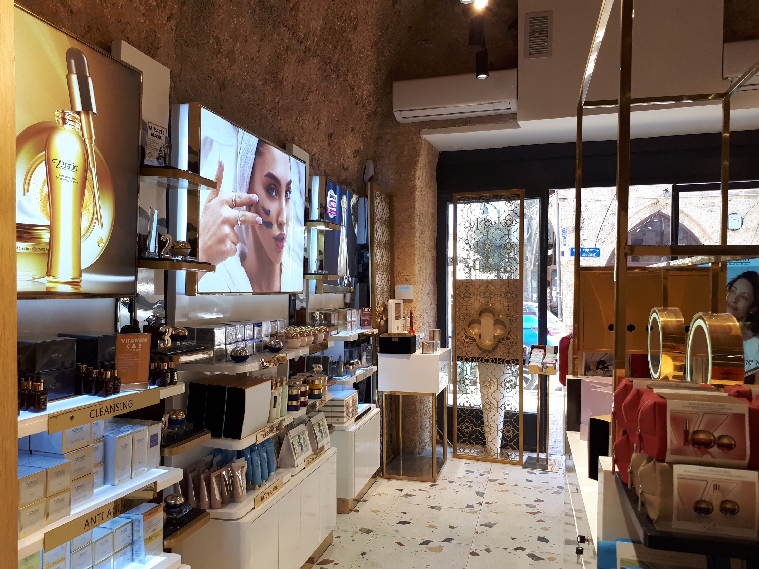 Innenansicht eines Kosmetikladens mit verschiedenen Produkten auf Gestellen, Bildschirmen an den Wänden, Deckenleuchten, einer Klimaanlage und Glastüren, die eine Person, ein Auto und ein Gebäude zeigen.