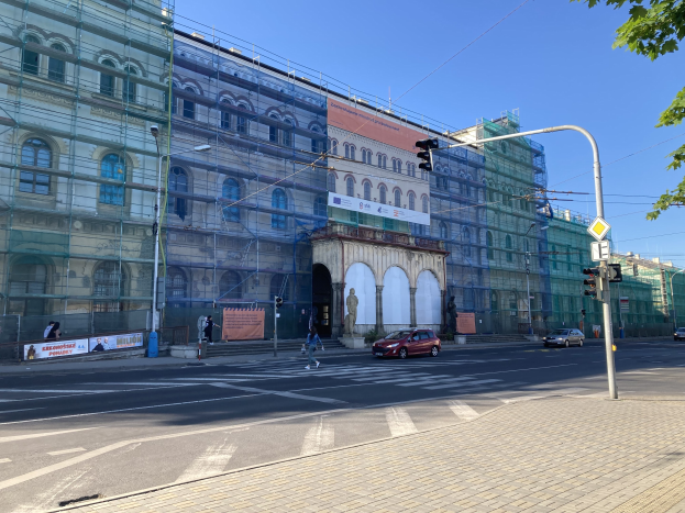 Ein großes Gebäude mit Gerüst, das renoviert wird, umgeben von Fahrzeugen, Fußgängern, Ampeln, Straßenschildern, Pfählen, Straßenlaternen, Strominfrastruktur, Bäumen und einem klaren blauen Himmel.