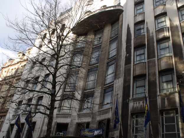 Gebäude mit Fenstern, einer Fahne und Flaggen an Stangen unten und einem Baum mit Ästen.