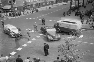 Eine Schwarz-Weiß-Szene eines Autounfalls an einer Straßenkreuzung, mit mehreren Fahrzeugen einschließlich eines Lieferwagens, einer Gruppe von Menschen darum und Gebäuden, Bäumen, Pfählen und Texttafeln im Hintergrund.